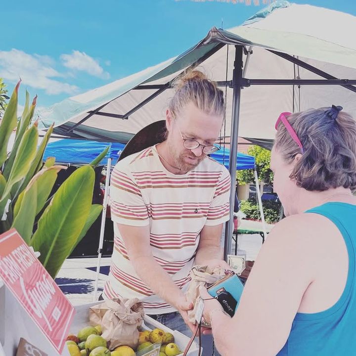 A man at the market booth handing a customer her purchase, a crate of guava beside them