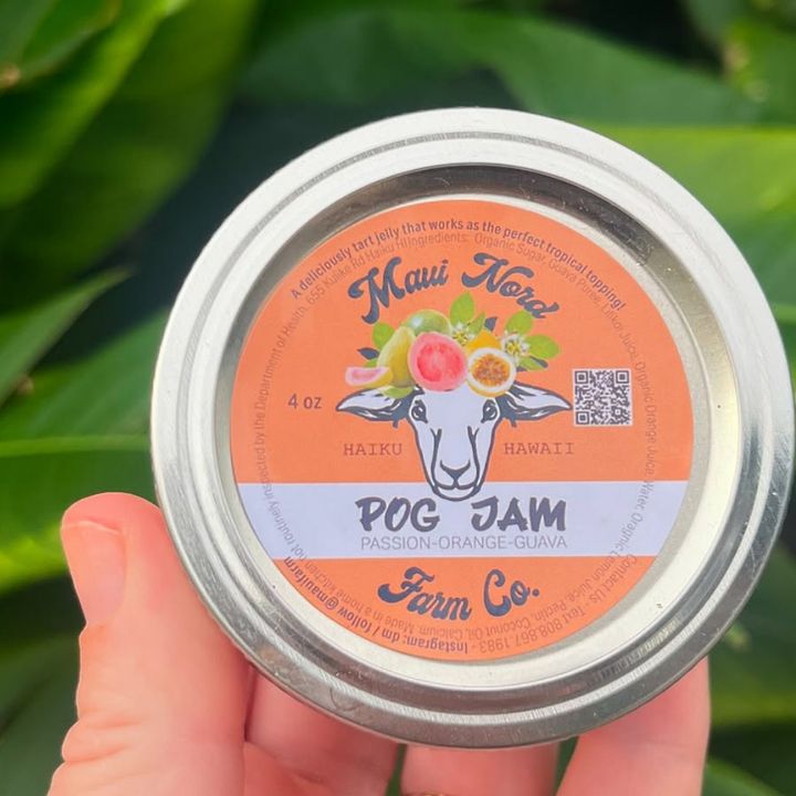 A hand holding a jar of POG jam against green leaves, the lid printed with the sheep mark