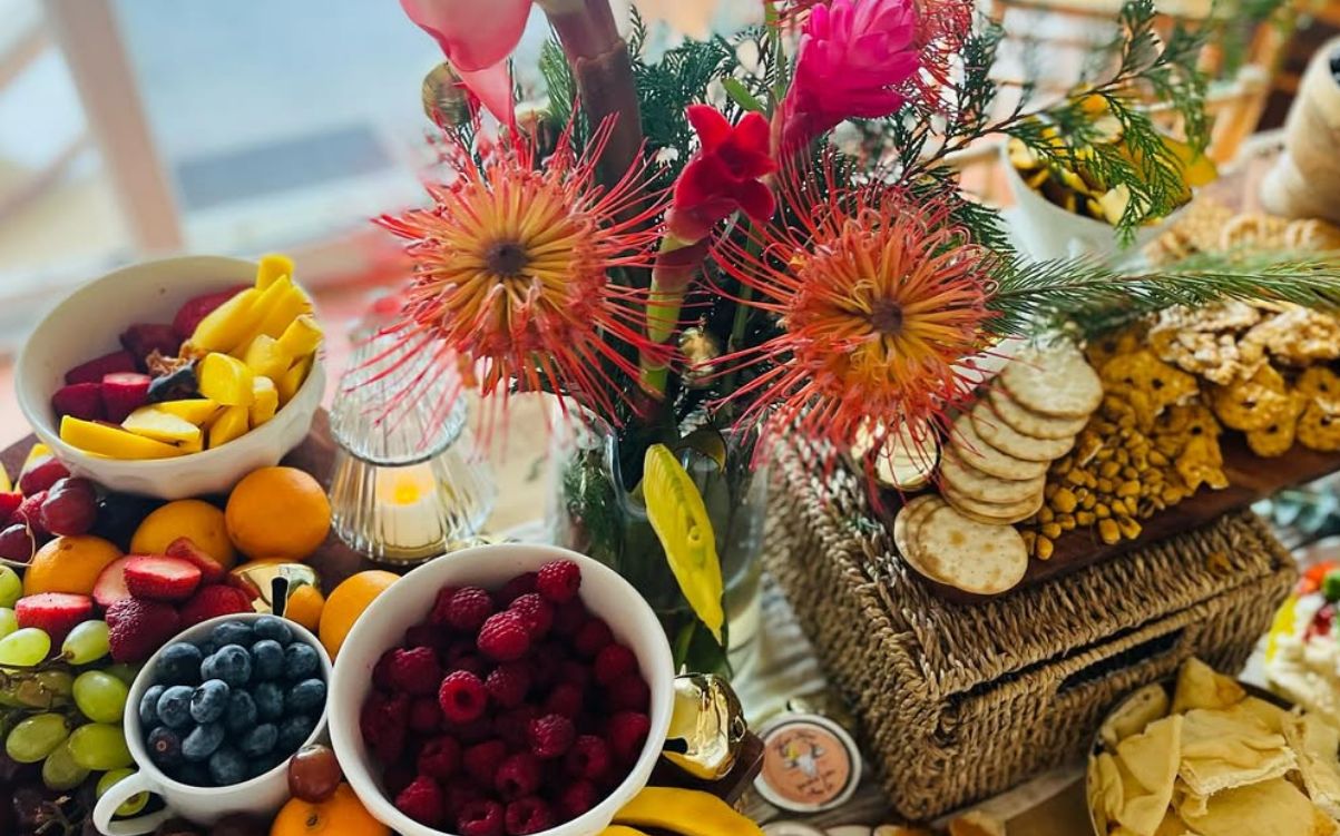 A table set with fruit, cheese and crackers on a floral tablecloth, with Maui Nord Farm jam jars at the front