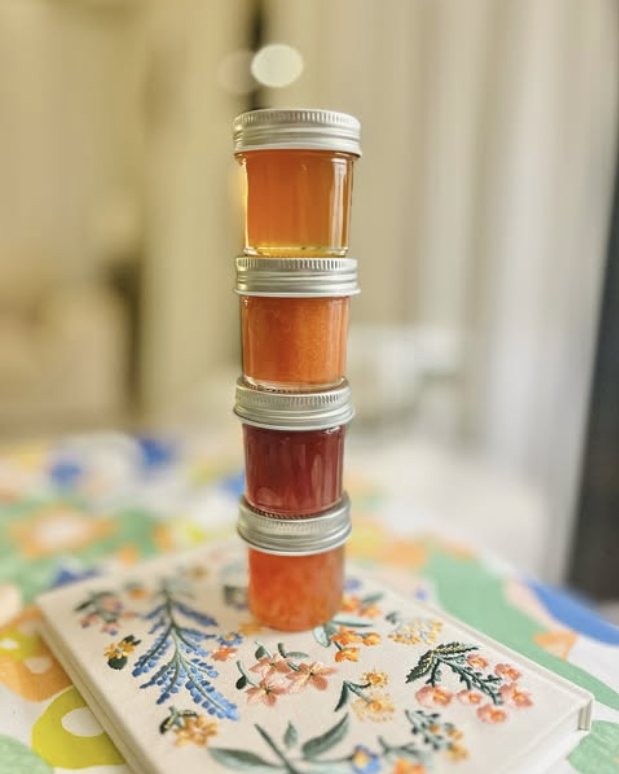 Four small jars of honey and jam stacked on a floral cloth, lit from behind